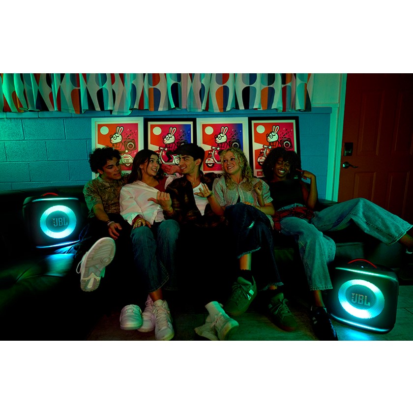 Group of people sitting on a couch with colorful posters and glowing speakers in the background.